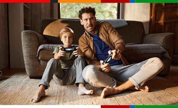 Mężczyzna i chłopiec siedzą na podłodze przed kanapą w salonie i grają razem w grę wideo, trzymając kontrolery do konsoli.